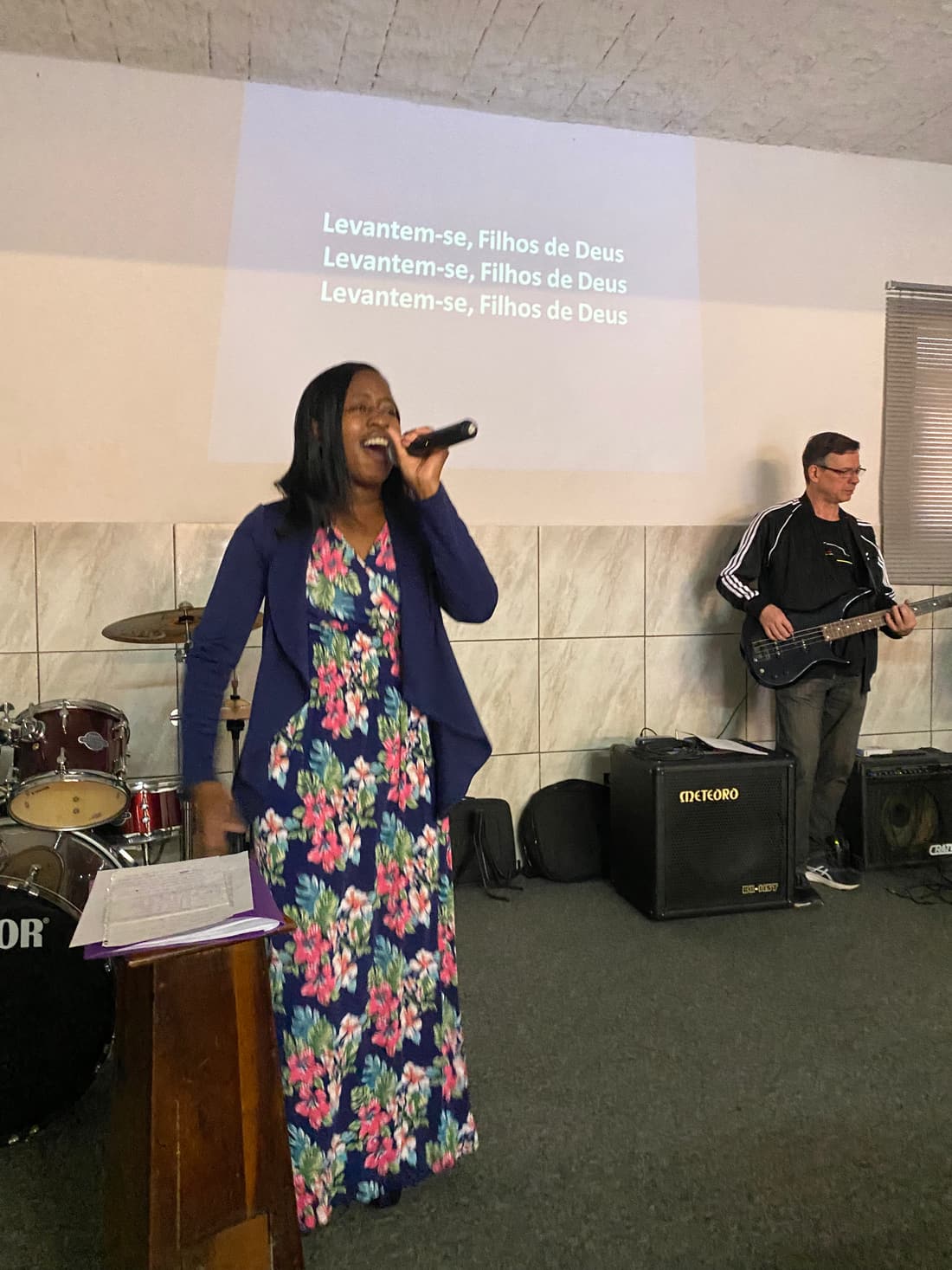 A vocalist singing with a bass player behind her, Portuguese lyrics on the wall